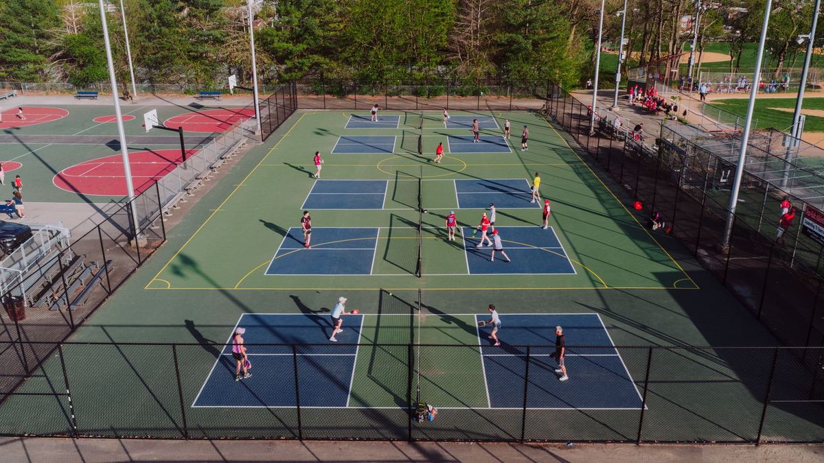 Outdoor pickleball courts seen from above, with players on several courts, ringed by trees